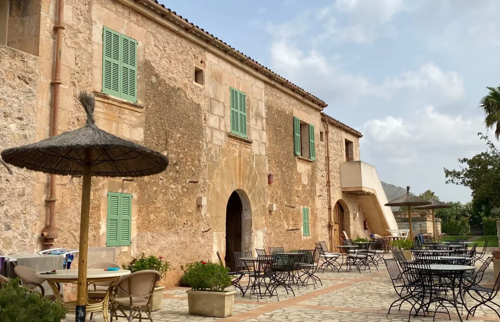 Terraza de una finca payesa en Mallorca con sillares de marés y contraventanas verdes, iluminada de lleno por el sol de frente, con mesas y un sombrajo de palma en el patio